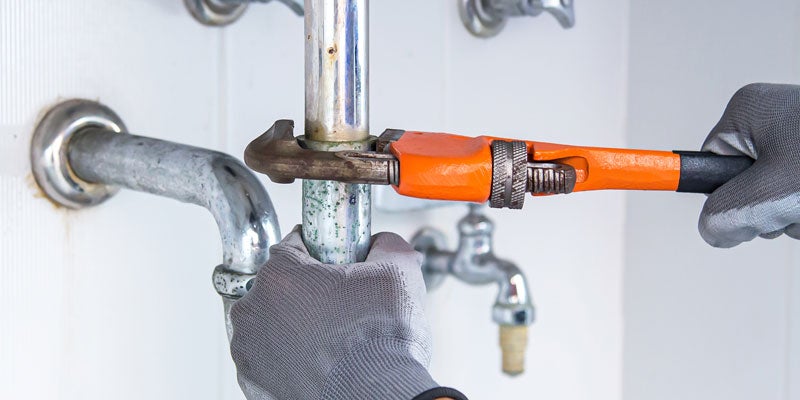 A plumber's gloved hands tightening pipes with an orange wrench