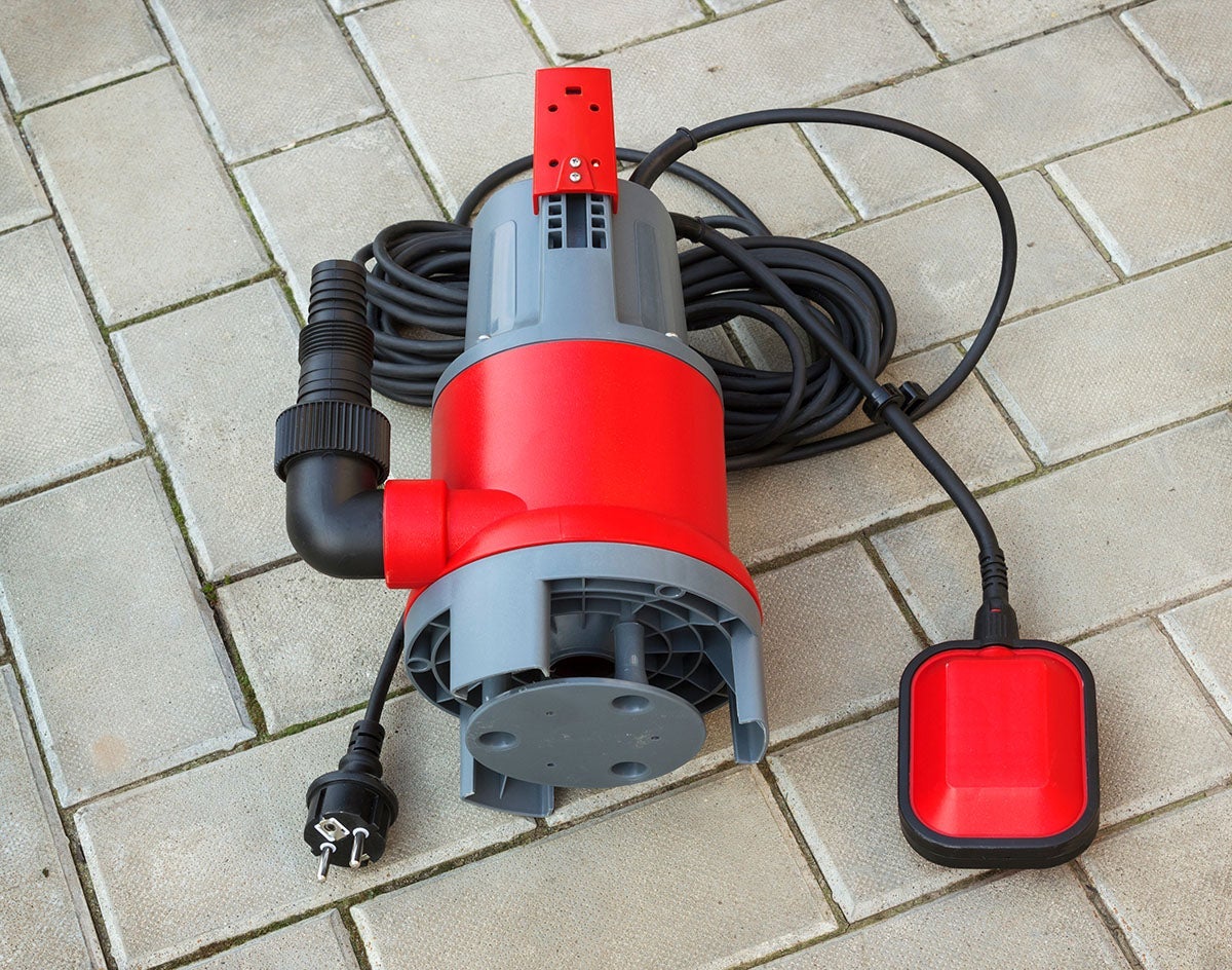 A red and grey submersible pump with a coiled power cord and a red float switch rests on concrete pavers.