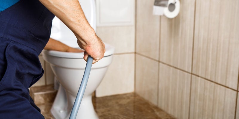 A person in blue overalls holds a drain snake, with a white toilet and tiled walls in the background.