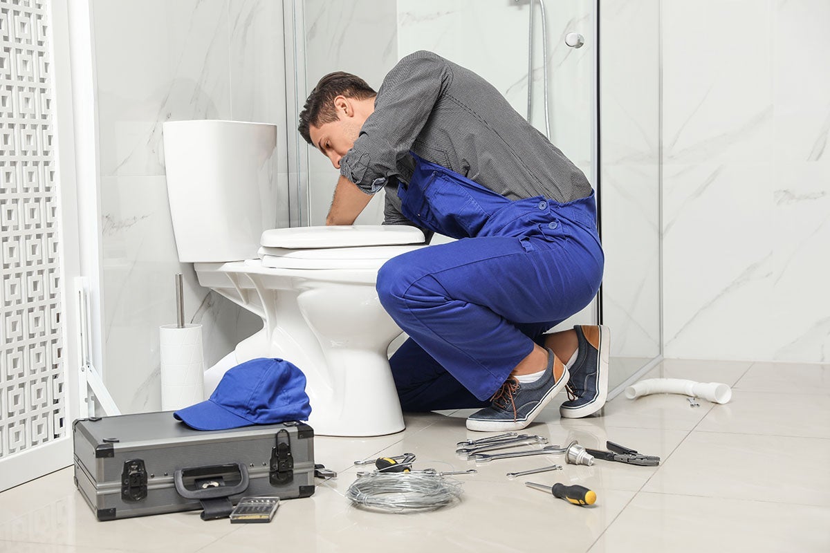 A male plumber in blue overalls installs or repairs a toilet in a modern white tiled bathroom.