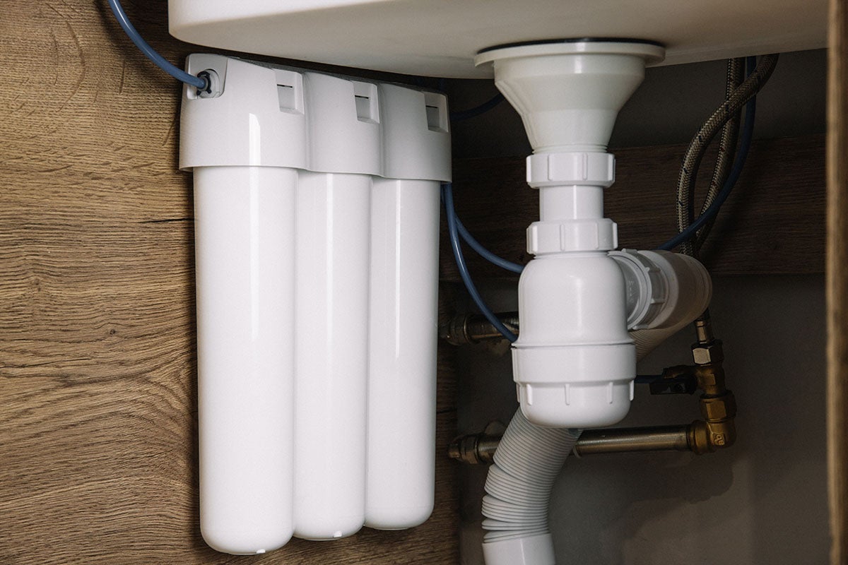 Under-sink water filter system with three white cartridges, next to white drainage pipes and brass plumbing.