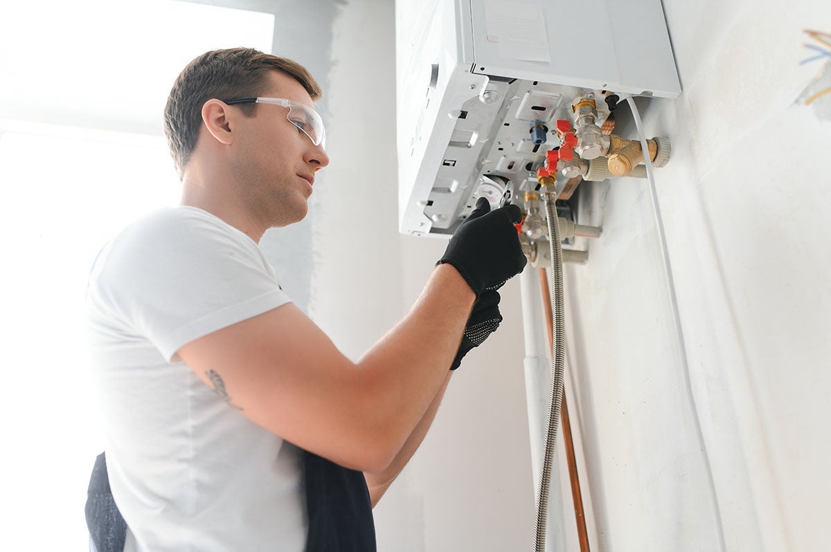 A man in safety glasses and work gloves adjusts the plumbing of a wall-mounted water heater or boiler.