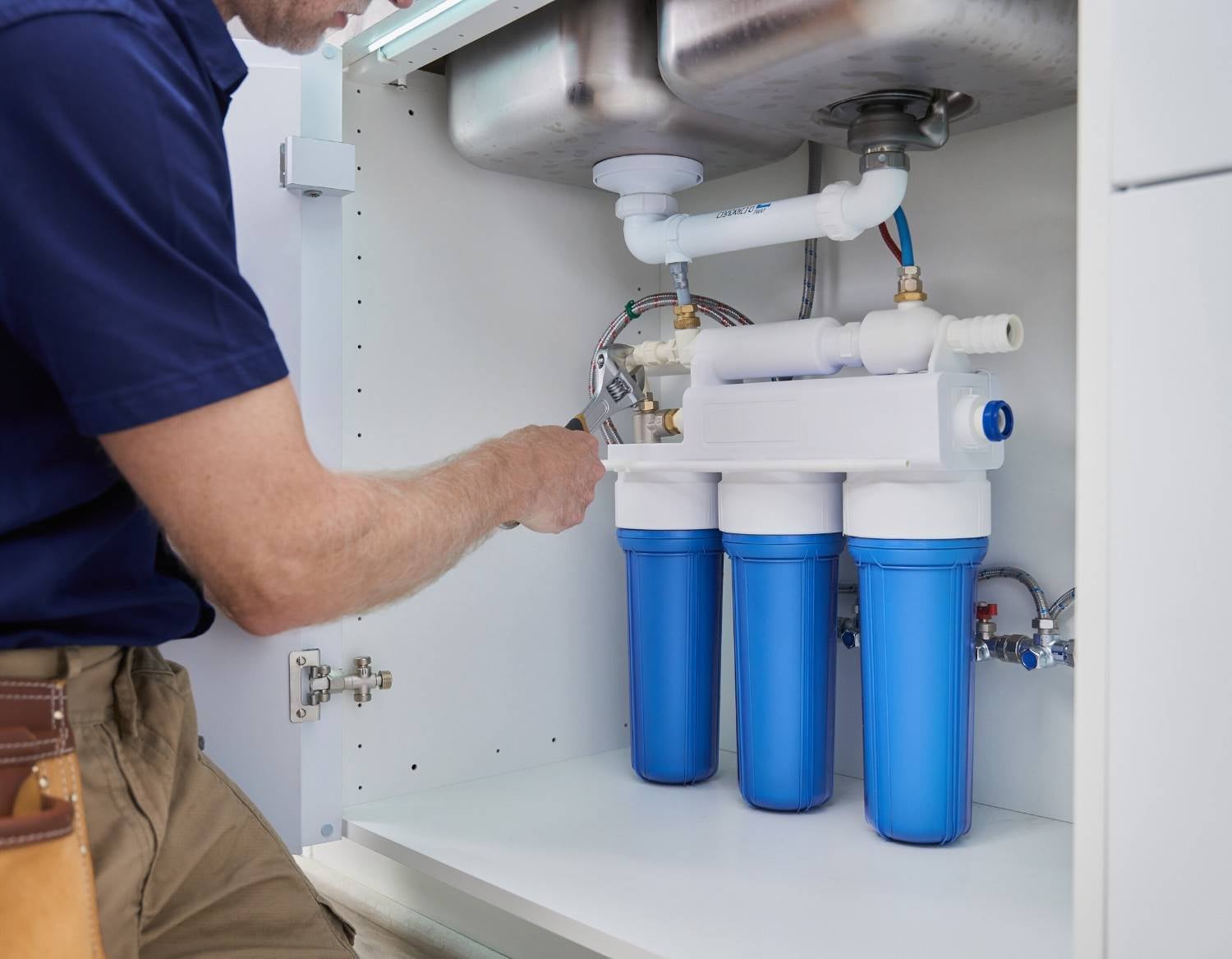A person tightens a pipe connection on a white water filter system with three blue cartridges under a sink.