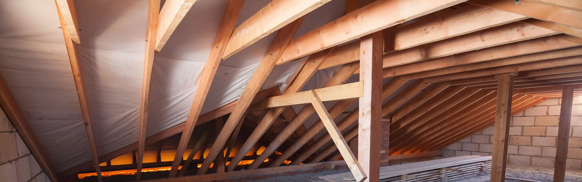Exposed wooden roof trusses and supports of an attic under construction, with a white vapor barrier.