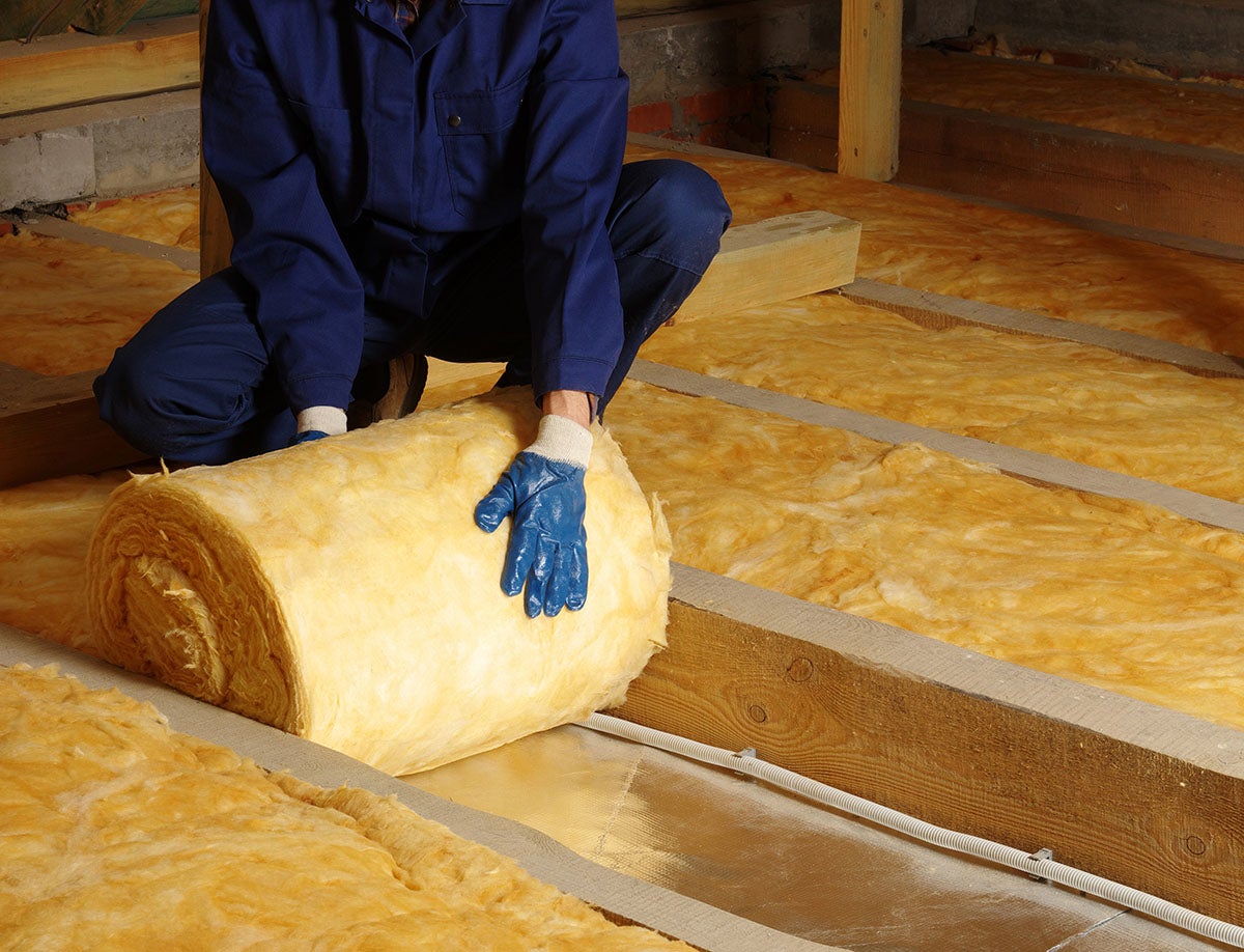 A person in blue overalls and gloves unrolls yellow fiberglass insulation over a subfloor between wooden joists.
